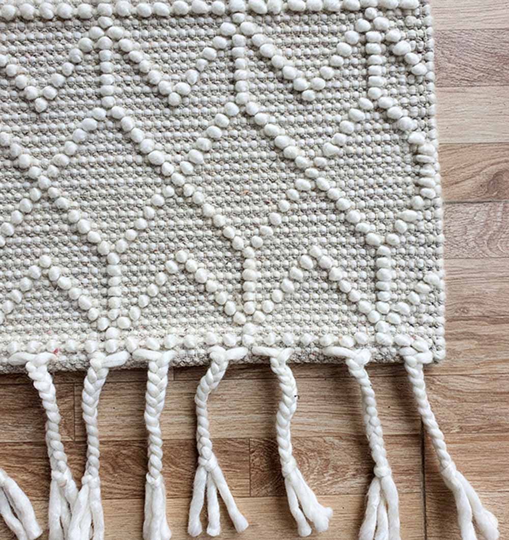 A close-up of the RAMSHA HOME Handmade Woven Rug (SRN-007) in natural white, with a raised geometric pattern and thick fringe, displayed on a wooden floor.