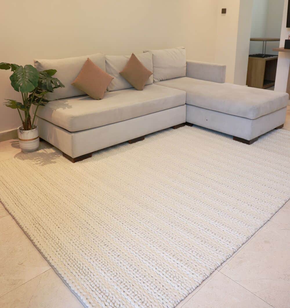 A modern living room features a light gray L-shaped sofa, two beige throw pillows, a potted green plant, and the Eid Festive Collection Natural White Rectangle Rug SRN 008—an elegant choice for Eid home décor.