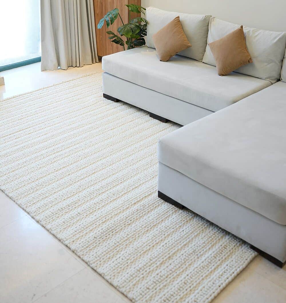 A modern living room with a light gray L-shaped sofa, two brown throw pillows, a plant, light curtains by the window, and the Eid Festive Collection Natural White Rectangle Rug SRN 008—perfect for displaying your Ramadan home décor.