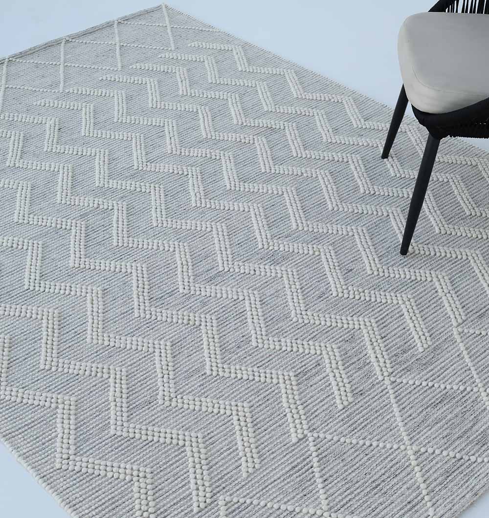 The Woven Rug Natural Color Perfect For Home Decor SRN-016 by RAMSHA HOME, featuring a textured zigzag pattern in gray and white, is partially covered by a modern chair with a light seat and dark frame.