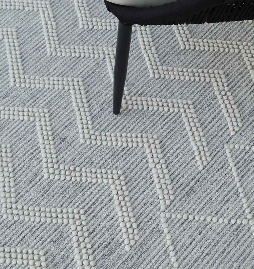 Close-up of the RAMSHA HOME Woven Rug Natural Color Perfect For Home Decor SRN-016, featuring a hand tufted wool texture in light gray with raised white zigzag tufts; a dark chair leg appears at the top.