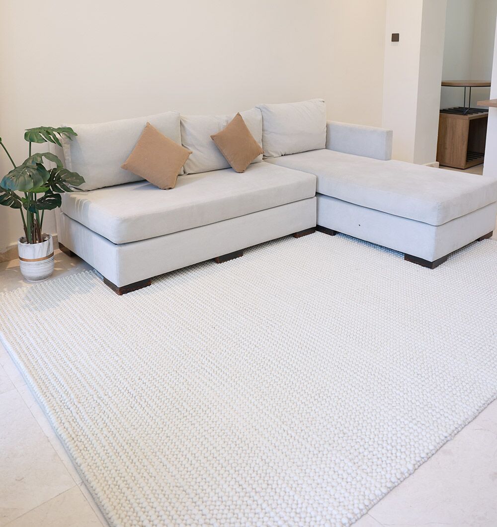 A light gray L-shaped sectional with beige and brown cushions sits on the RAMSHA HOME Natural Woven Rug For Home Decor SRN-026. A potted plant stands beside the sofa in a bright, minimalist living room.