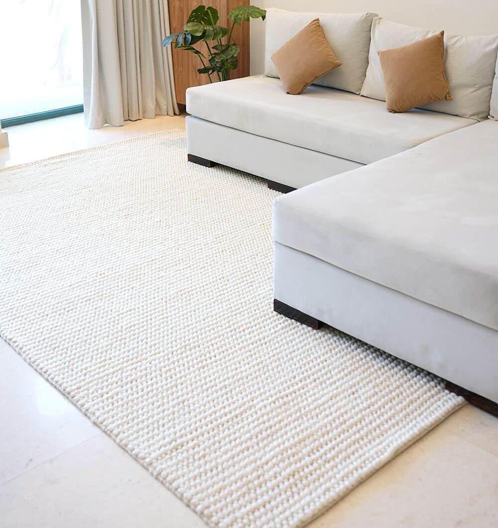 A modern living room featuring the RAMSHA HOME Natural Woven Rug For Home Decor SRN-026, a light gray sectional with beige and brown pillows, plus a green potted plant by a window with sheer curtains.