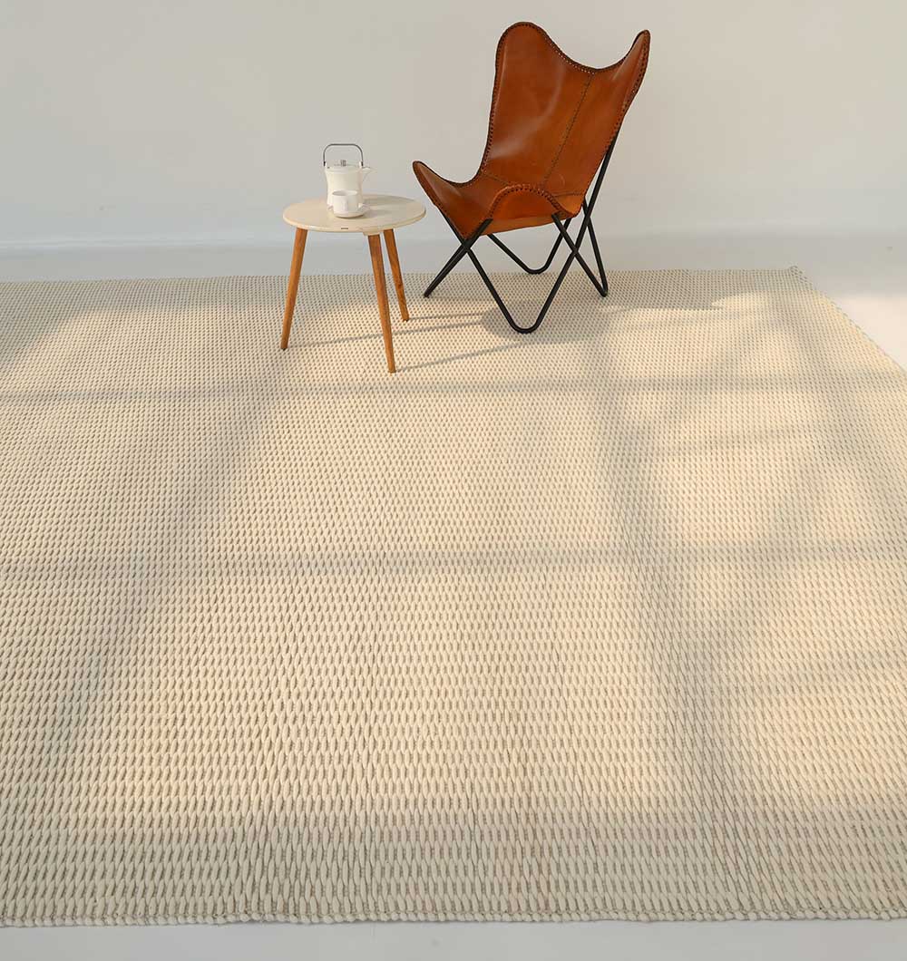 A minimalist room features the RAMSHA HOME Handmade Natural White Woven Rug SRN-030, a brown leather butterfly chair, and a small round wooden side table with a glass teapot and cup. Soft sunlight casts gentle shadows across the floor.