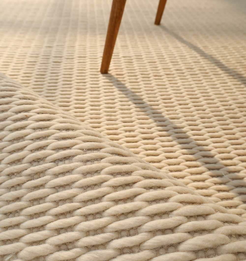 Close-up of the Handmade Natural White Woven Rug For Home Decor SRN-030 by RAMSHA HOME, showcasing textured natural fibers and thick braided loops, with a wooden furniture leg's shadow adding warmth to the scene.