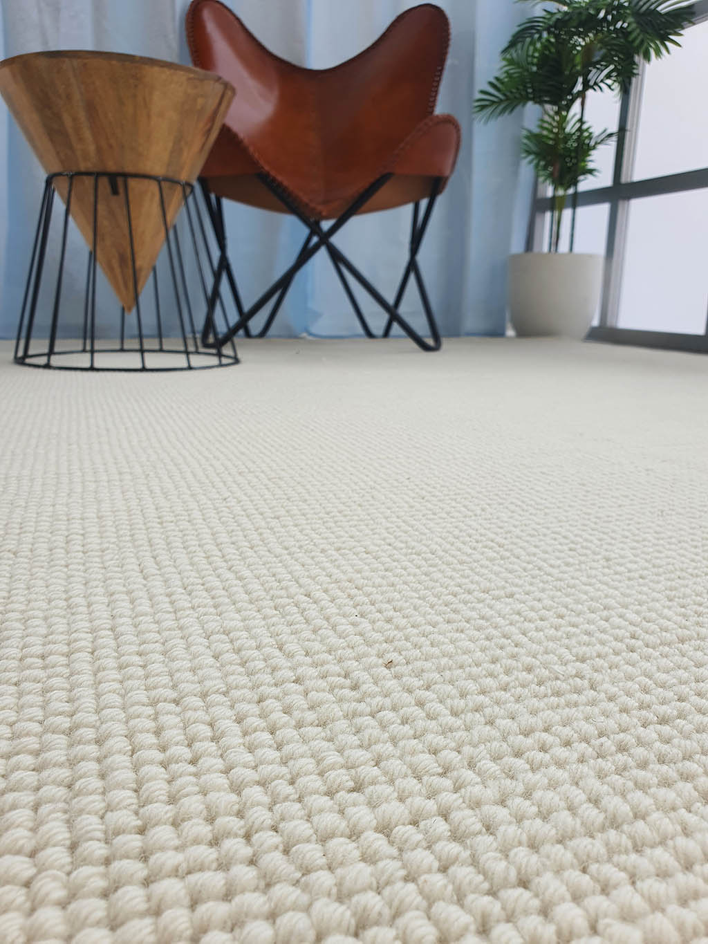 Close-up of the Solid Loop Pile Woven Rug For Home Decor SRN-032 in beige, placed in a modern room with a brown leather chair, wooden side table, potted plant, and large windows with sheer curtains.