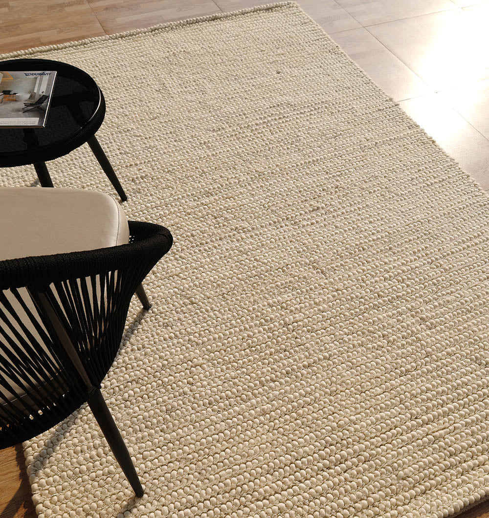 A Handmade Natural Color Woven Rug For Living Room SRN-036 by RAMSHA HOME in cream is spread on a sunlit floor, next to part of a black and white chair and a black round side table with a magazine in the corner.
