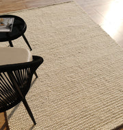 A Handmade Natural Color Woven Rug For Living Room SRN-036 by RAMSHA HOME in cream is spread on a sunlit floor, next to part of a black and white chair and a black round side table with a magazine in the corner.