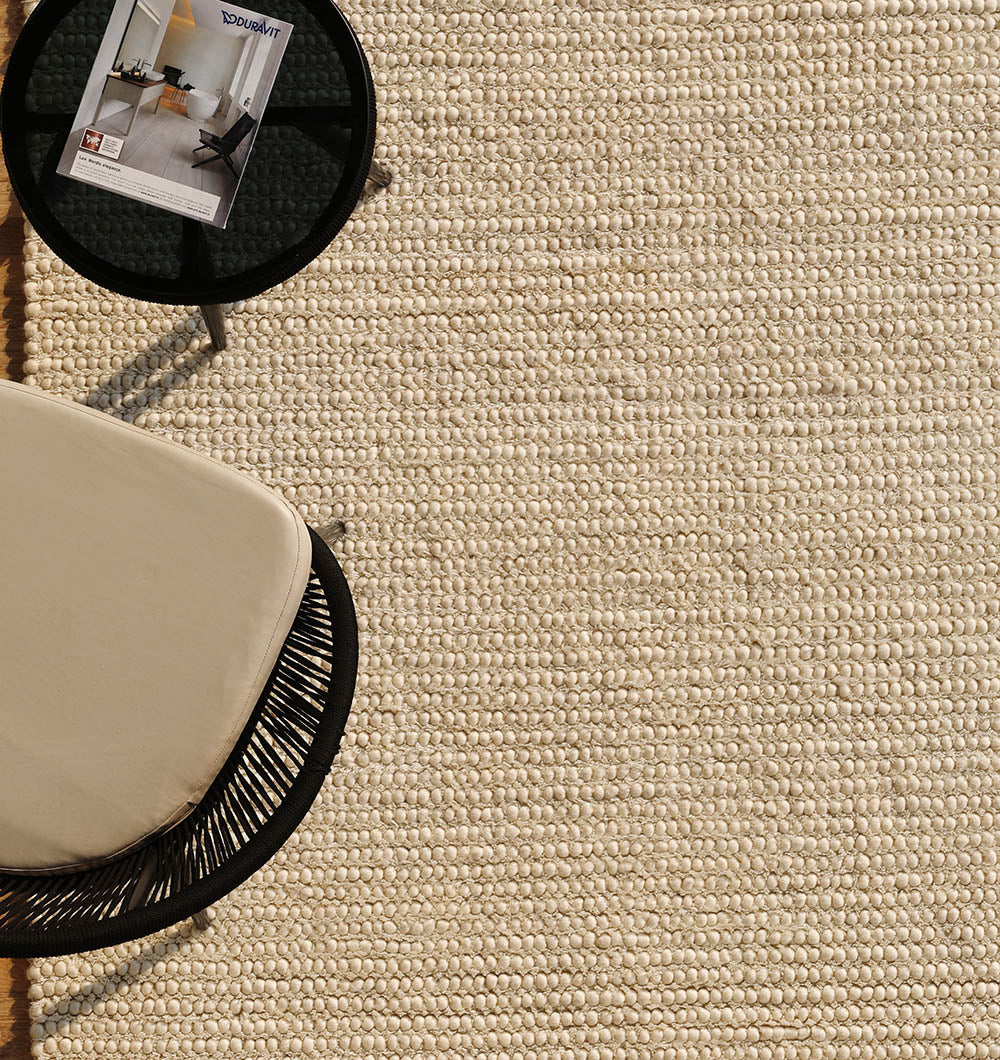 A top view of the RAMSHA HOME Handmade Natural Color Woven Rug For Living Room SRN-036, paired with a black round side table holding a magazine and part of a modern chair featuring a beige cushion.