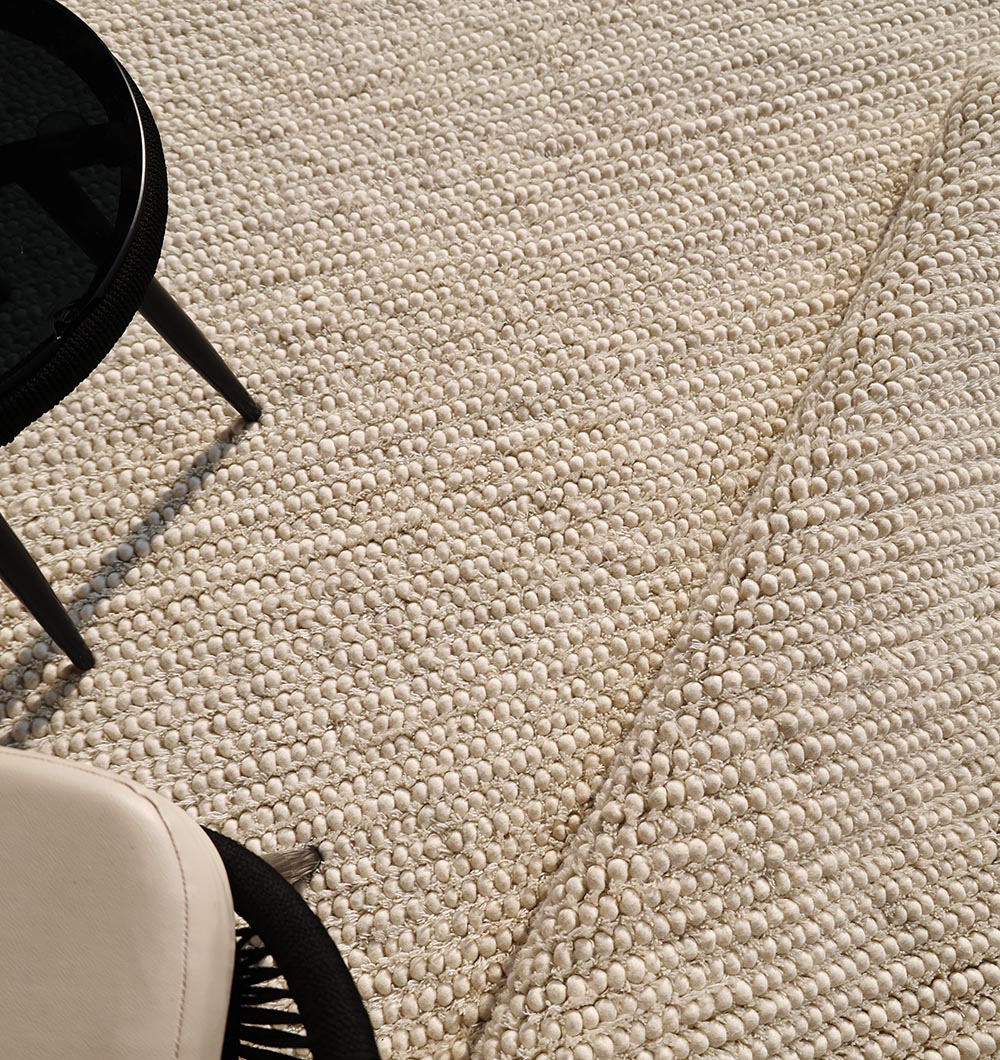 A close-up of the RAMSHA HOME Handmade Natural Color Woven Rug For Living Room SRN-036 with textured off-white fibers and a slight fold. A round black table and white chair appear in the lower left corner.
