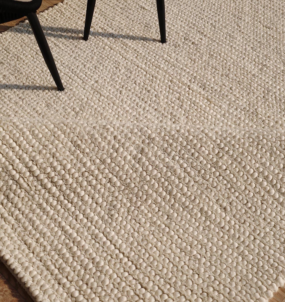 The RAMSHA HOME Handmade Natural Color Woven Rug For Living Room SRN-036 features a textured, bumpy pattern and is shown partially draped under two black chairs on a wooden floor.