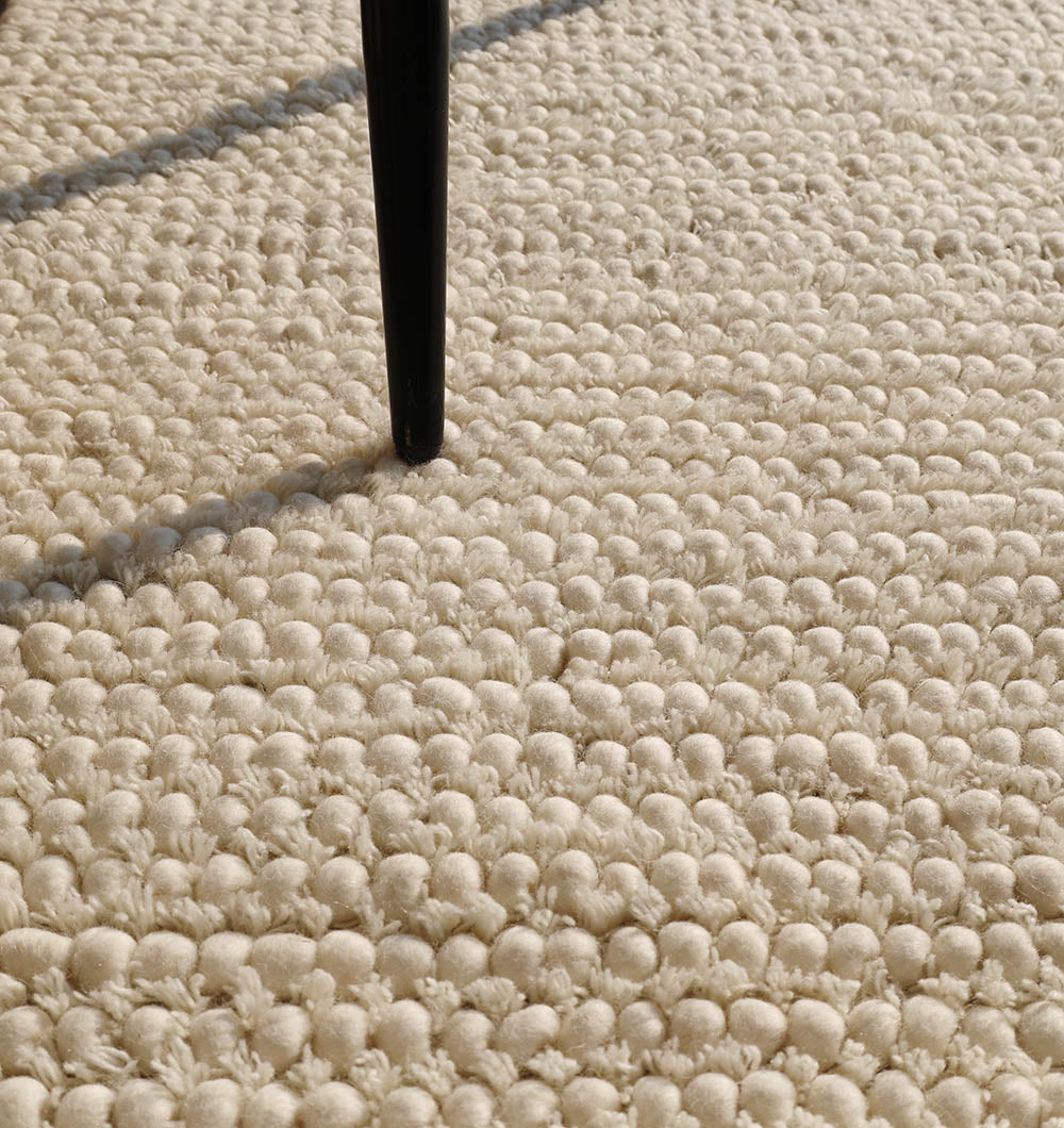 Close-up of the RAMSHA HOME Handmade Natural Color Woven Rug For Living Room SRN-036, showing its textured cream surface with a black furniture leg casting a shadow on the soft weave.