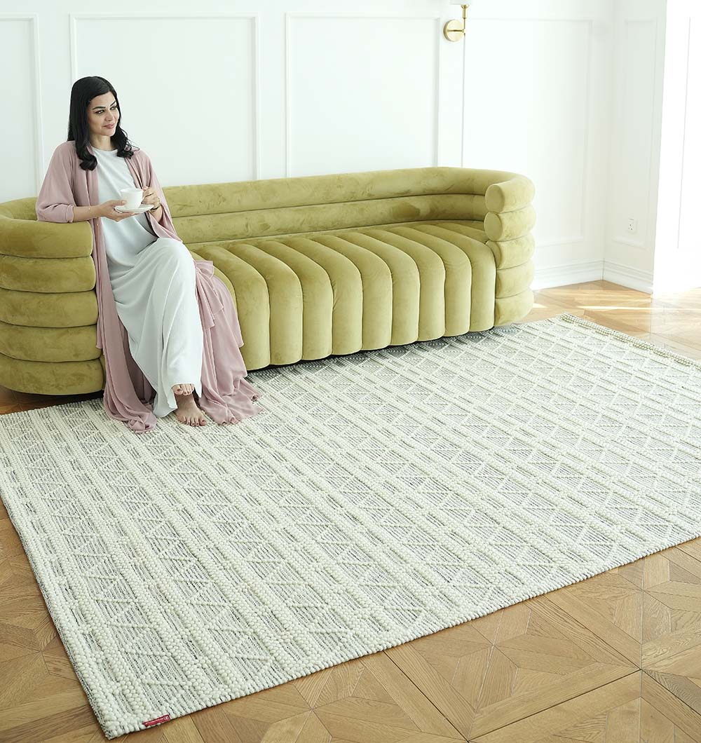 A woman in a light dress and pink robe sits barefoot on a mustard yellow sofa, holding a cup. The room features the RAMSHA HOME Wool Woven Rug Area Rug with Natural Color SRN-040 over parquet wood floors and crisp white walls.