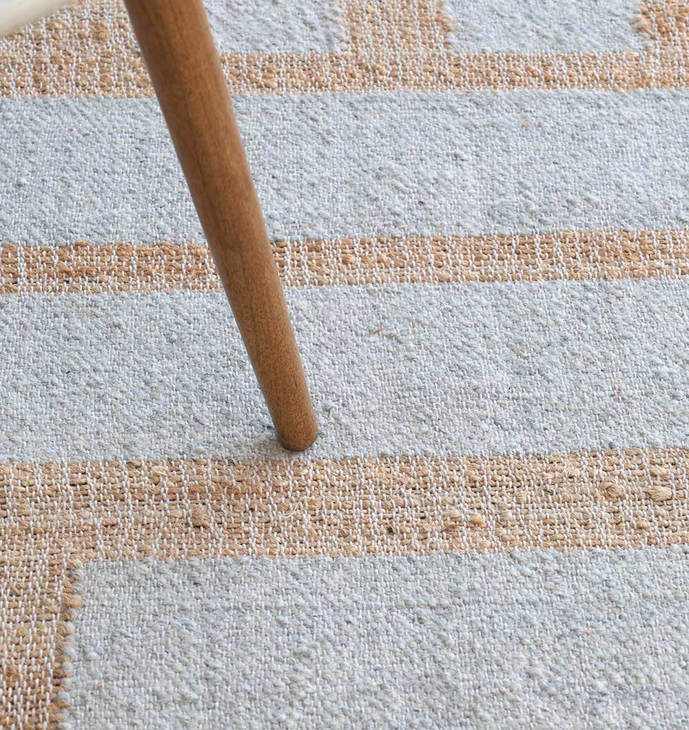 A wooden chair leg sits on the RAMSHA HOME Jute & Wool Natural Color Hand Woven Rug WR-036, featuring a flat weave with a light blue and beige checkered pattern.