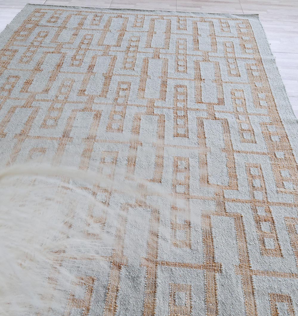 The RAMSHA HOME Hand Woven Decor Area Rug WR-037 features a geometric light brown pattern on a light beige background, adding interest to a light wood floor. Decorative dried grass is slightly visible in the lower left corner.