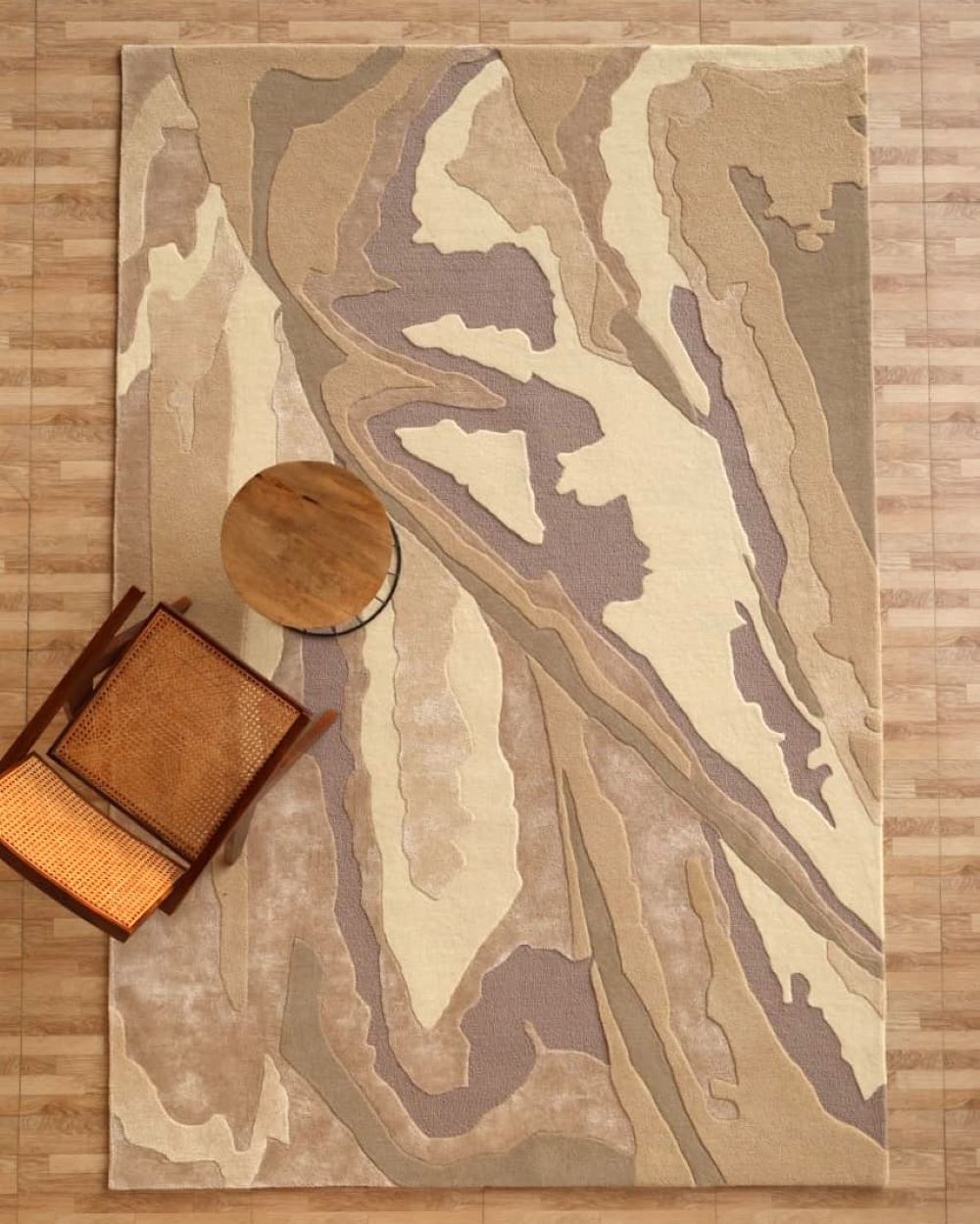 The RAMSHA HOME Abstract Wool & Viscose Handmade Carpet 200x300 – HTWV-179, featuring taupe and earth-toned abstract shapes, lies on a wood floor beside a woven-seat chair and round table.