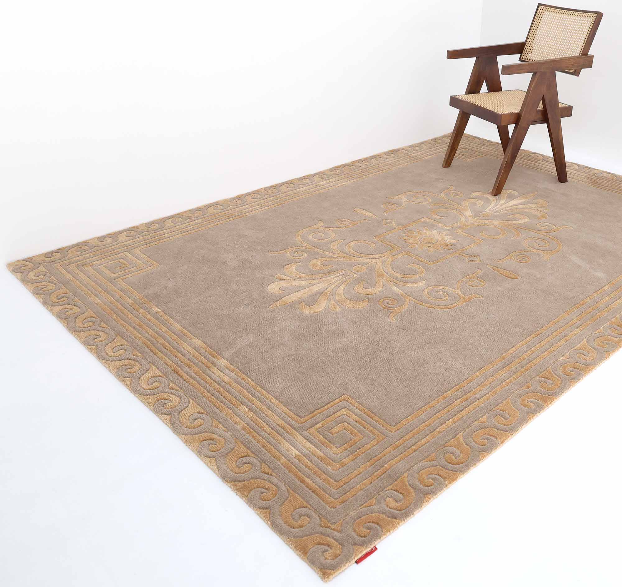 A Beige Rectangle Novara Rugs- CMN 22 (200x300cm) by RAMSHA HOME, featuring intricate gold patterns and a Greek key border, lies on a white floor with a wooden chair beside it.